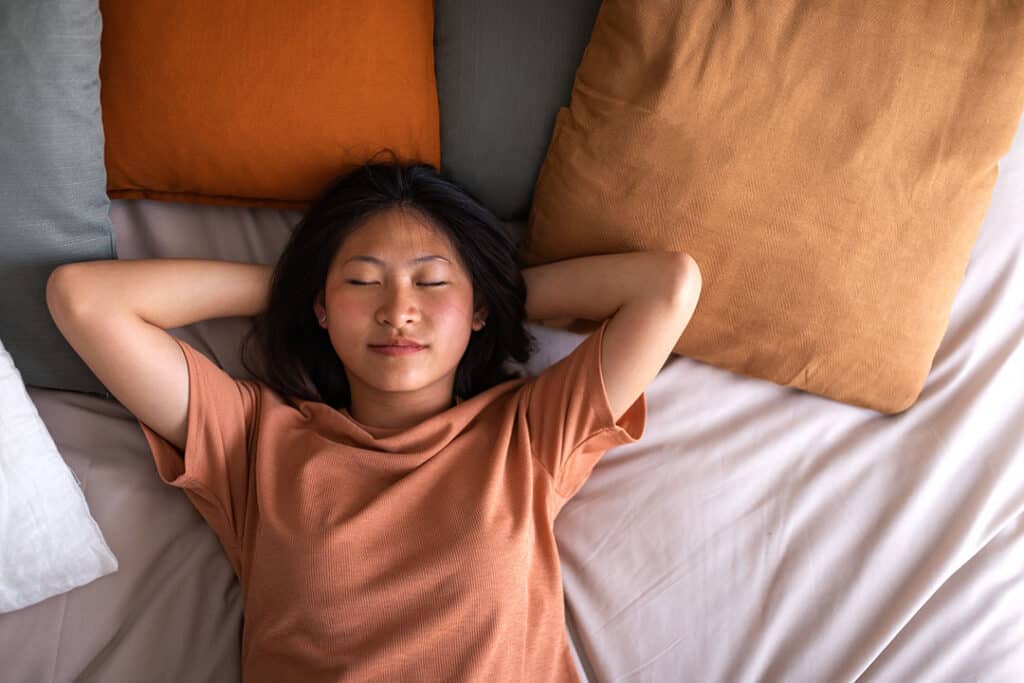 A person with dark hair lies on a bed with eyes closed, hands behind their head, possibly resting after a bout of whiplash, surrounded by orange and gray pillows.