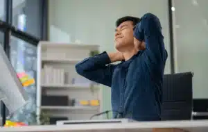 A man sits at a desk in an office, leaning back in his chair with his hands on his neck, appearing to stretch or relieve recurring neck pain.