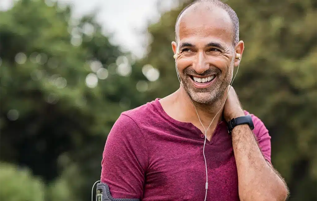 A man wearing a maroon shirt and headphones smiles outdoors, with one hand behind his neck—possibly easing recurring neck pain—with trees in the background.