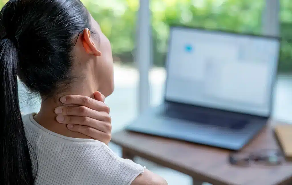 A woman sits at a desk with a laptop, holding her neck as if experiencing recurring neck pain or discomfort.