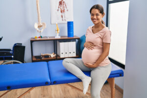 A pregnant woman sits on a blue examination table in a medical office, smiling and holding her belly, possibly preparing for the Webster Technique.