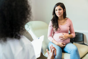 A pregnant woman sits in a chair, holding her belly and listening as a Webster Certified Chiropractor offers guidance during a consultation in a medical office.