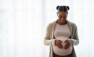A pregnant woman stands indoors, smiling and forming a heart shape with her hands over her belly. She is wearing a light sweater and hoop earrings.