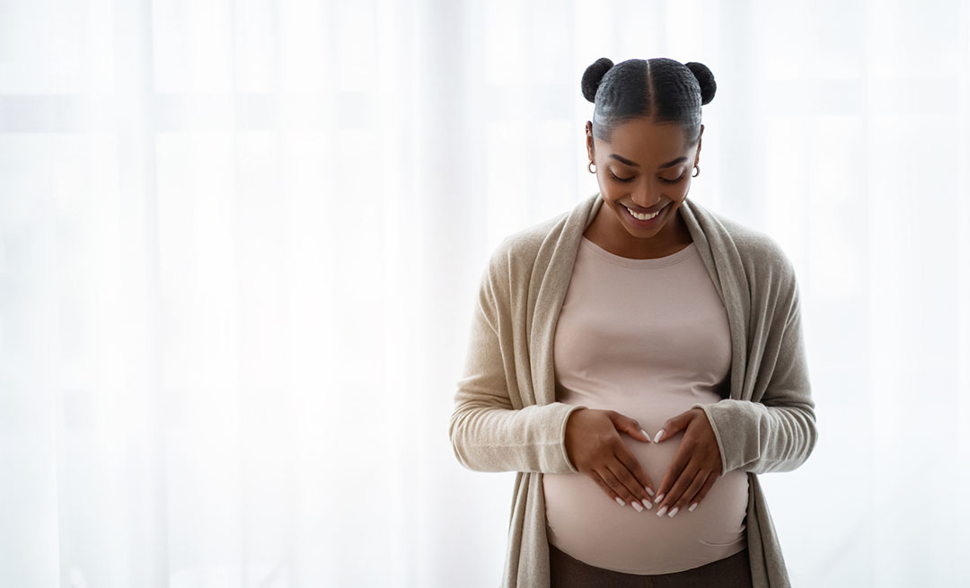 A pregnant woman stands indoors, smiling and forming a heart shape with her hands over her belly. She is wearing a light sweater and hoop earrings.