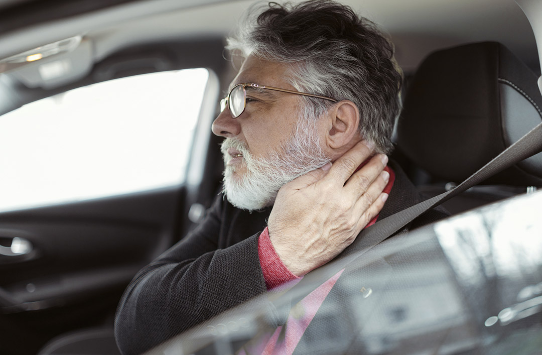 An older man with glasses sitting in a car driver's seat touches his neck, appearing to be in discomfort or pain, possibly experiencing delayed symptoms.