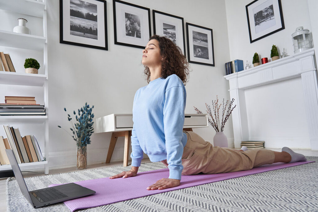 Woman with fibromyalgia practicing yoga in a living room, performing an upward facing dog pose on a purple mat while looking at a laptop.