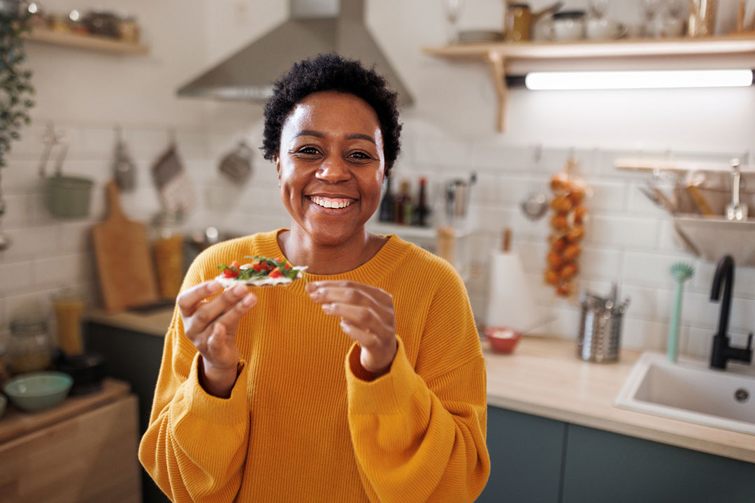 A person in a yellow sweater, living with fibromyalgia, smiles while holding a slice of food in a modern kitchen with white tiles and wooden shelves in the background.