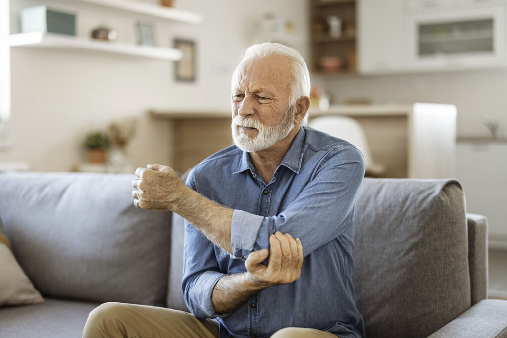 An older man sits on a couch, holding his elbow with a pained expression, suggesting discomfort or pain in his arm—possibly due to fibromyalgia.